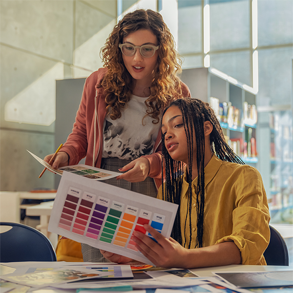 JFM Technology Two women collaborate at a table reviewing color swatches from a printed palette in a bright library or classroom
